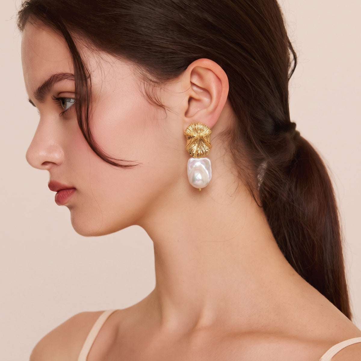 Woman wearing a gold petal-shaped stud earring with a baroque pearl drop, side profile