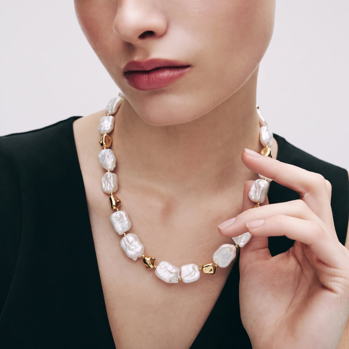 Woman wearing a necklace with square baroque pearls and gold stone-shaped links, hand touching necklace