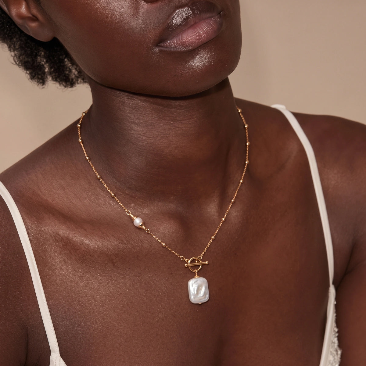 Woman wearing a thin gold chain necklace with a small pearl accent and a rectangular pearl pendant